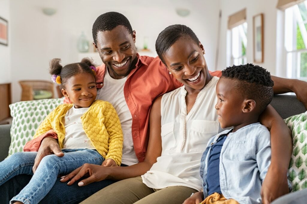 Happy Black family enjoying quality time together at home, featuring parents and two children, emphasizing comfort and family bonding in a cozy living room setting.