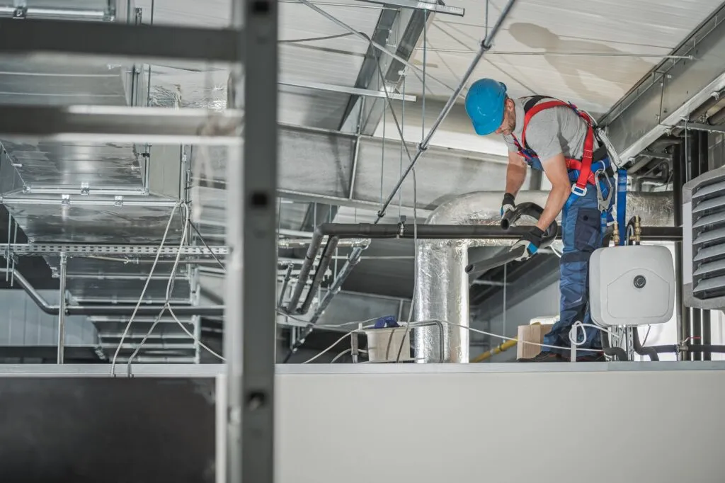 HVAC technician working on system assembly inside a commercial building, featuring ductwork and safety gear, emphasizing professional installation services for efficient heating and cooling systems.