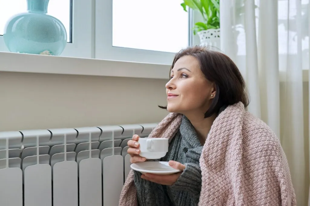 Woman wrapped in a cozy blanket, sitting near a home heating radiator, holding a cup of tea, enjoying warmth during winter in a well-lit room.