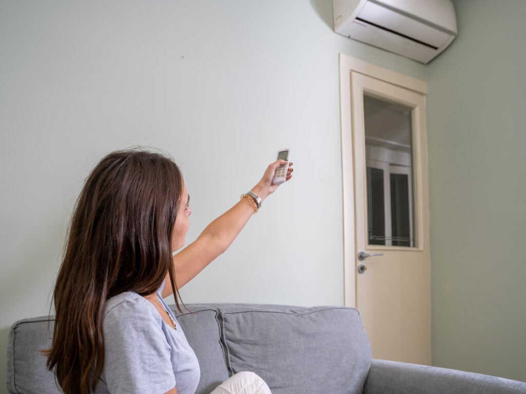 Woman sitting on a sofa using a remote control to operate a ductless mini-split air conditioning unit on the wall, illustrating home comfort and HVAC efficiency.