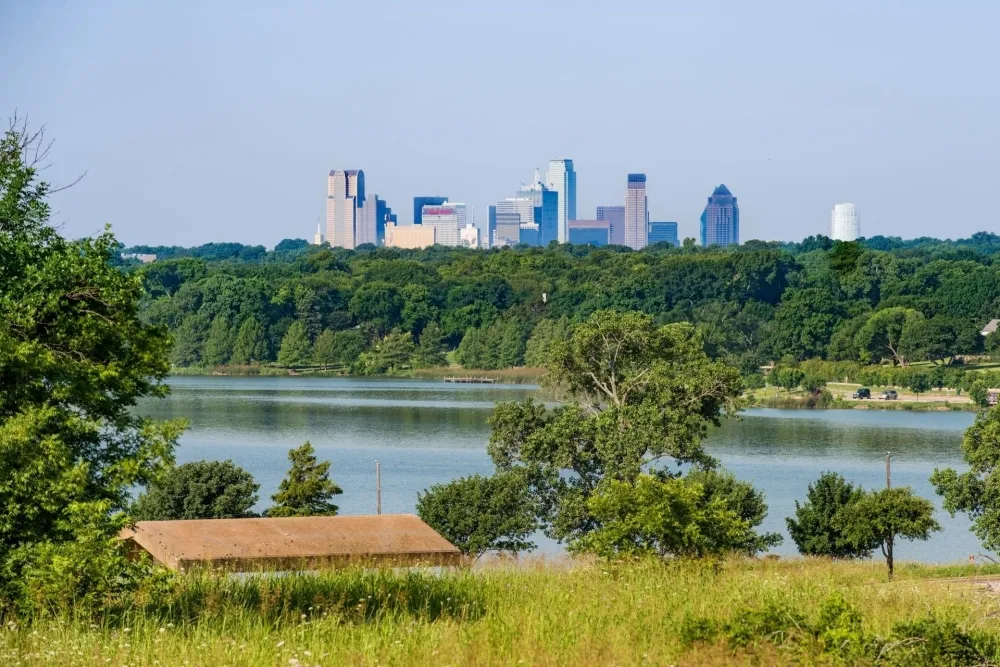 Scenic Dallas, TX skyline view across a lake, framed by lush trees and greenery. Consider ductless heating for your home's comfort needs.