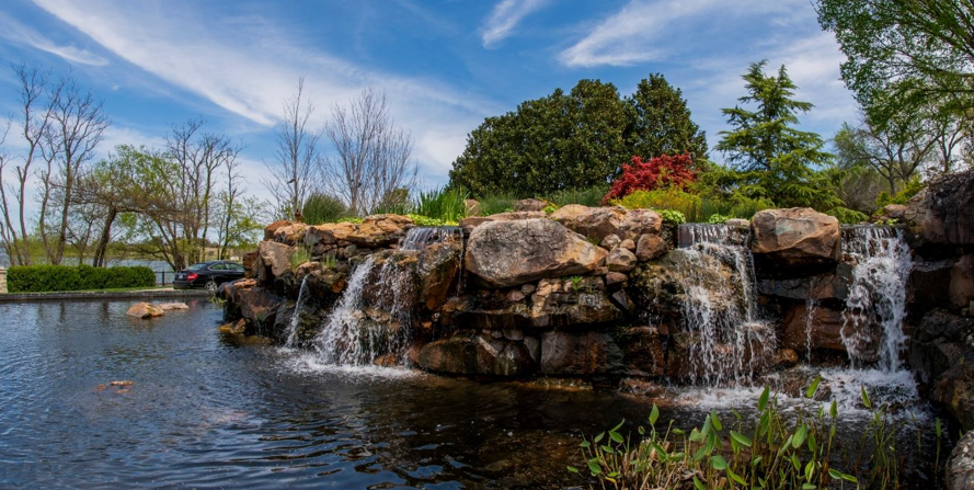 Scenic view of a landscaped pond with a waterfall, featuring lush greenery and colorful flowers, set against a clear blue sky, relevant to enhancing home comfort through air balancing in Dallas.