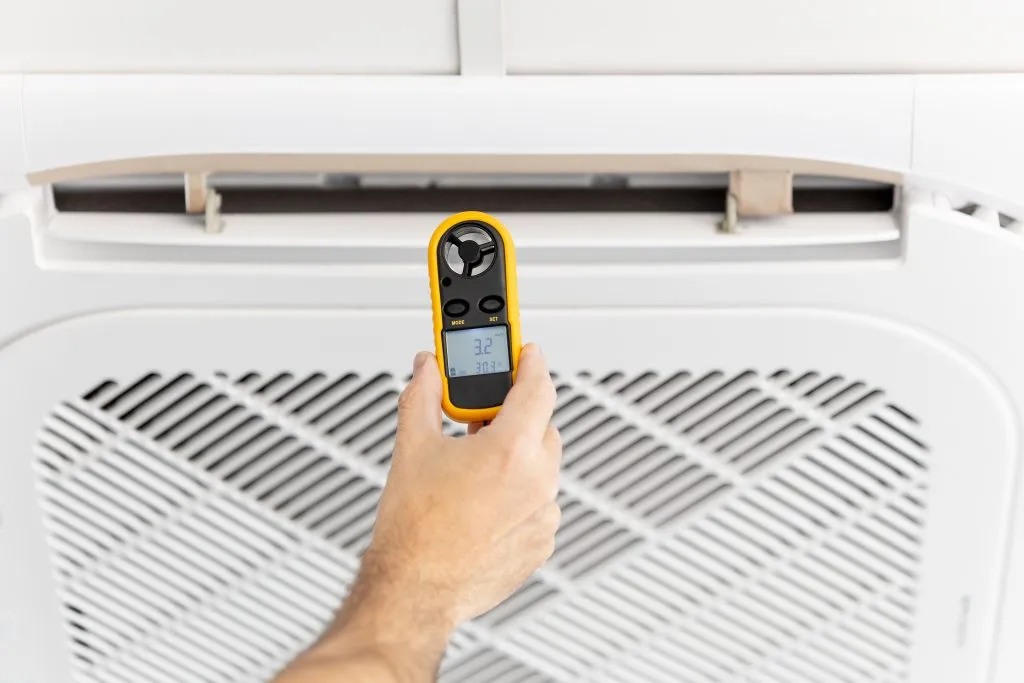 IAQ assessment: Hand holding anemometer measuring airflow in ceiling vent, ensuring optimal indoor air quality and ventilation performance.